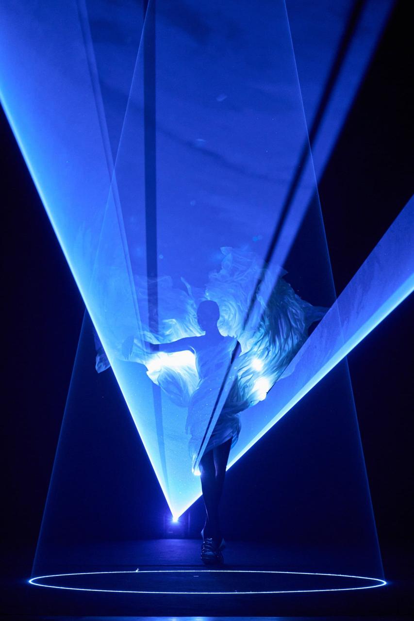 A performer on a circular stage, surrounded by luminous fabric and blue laser beams